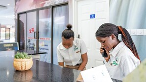 A reception desk with a potted plant, a white sign with a logo, and a glass door in the background. - The Shrub lodge (Victoria Falls)