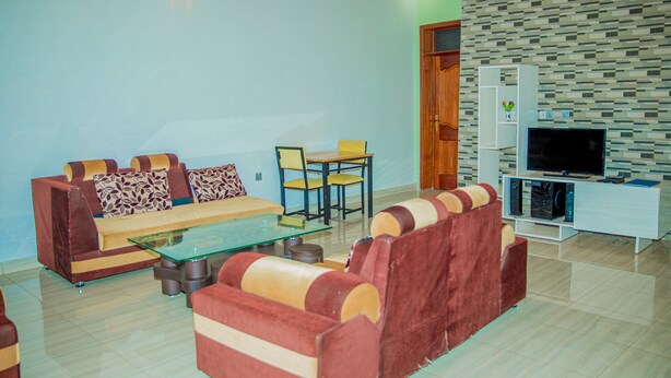 A living room with a sofa set, a glass coffee table, a TV stand with a flat-screen TV, and a patterned wall. -  LEISURE INN (Muhoza)