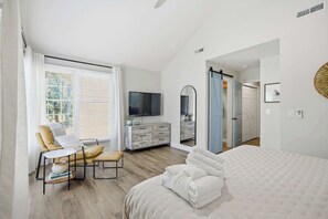 A bedroom with a bed, a TV, a desk with a chair, a dresser, and a mirror.