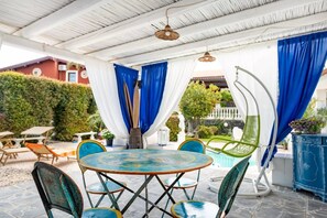 Ein überdachter Außenbereich mit einem runden Tisch, Stühlen, einem Pool und einem weißen Zaun im Hintergrund.