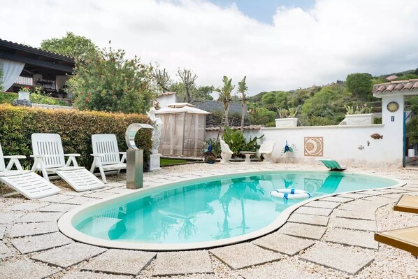 Ein Pool mit Liegestühlen, einer Pergola und einem Garten mit verschiedenen Pflanzen und einem kleinen Gebäude.