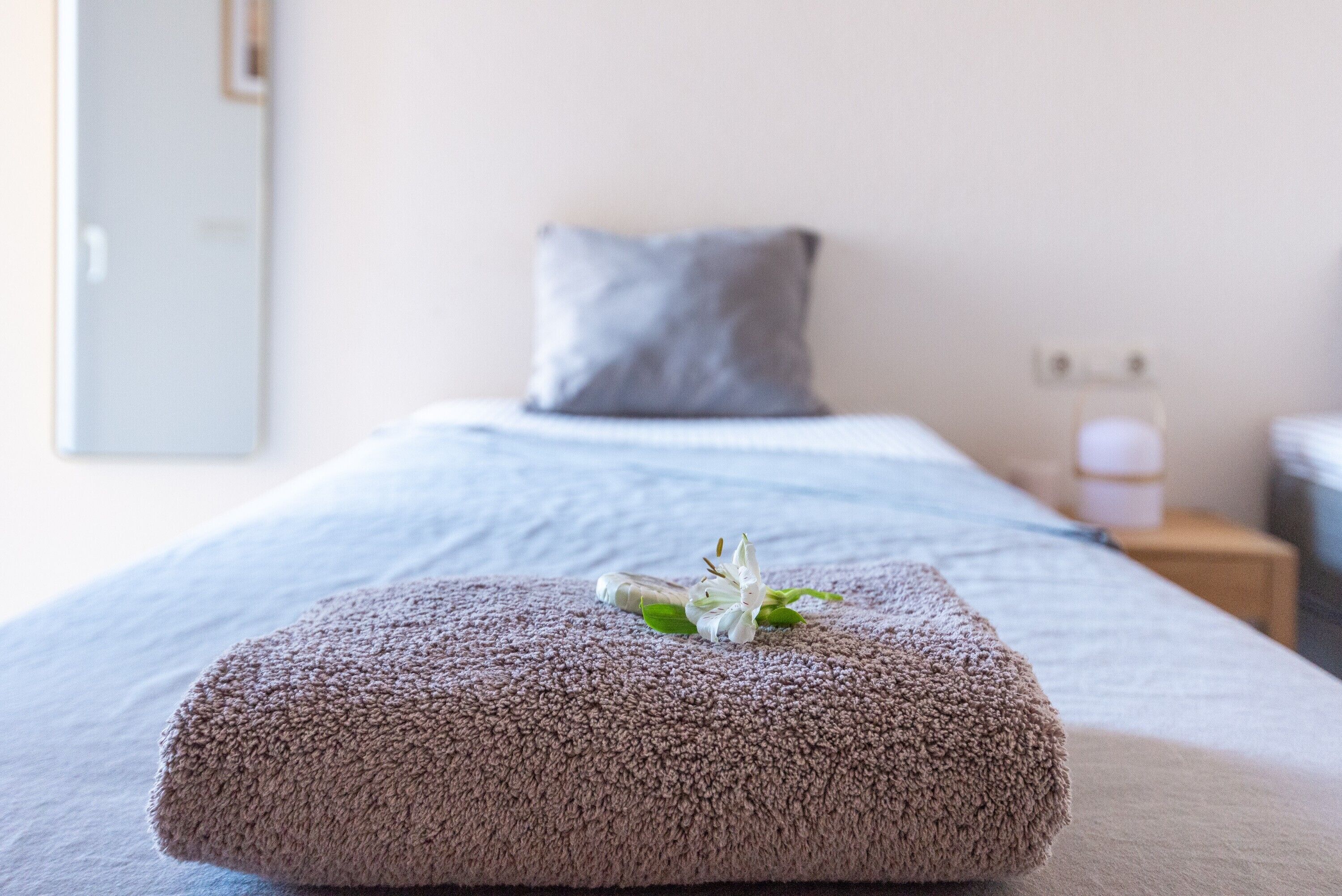 Ein ordentlich bezogenes Bett mit einem gefalteten Handtuch und einer weißen Blüte darauf, ein graues Kopfkissen und ein Nachttisch mit einer Lampe.