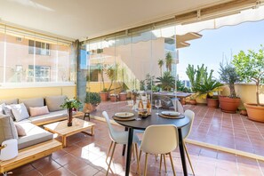 Une terrasse extérieure avec une table à manger, des chaises et un canapé. On y trouve des plantes en pot et, en arrière-plan, une vue sur des bâtiments.
