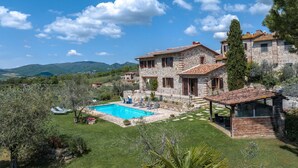Una villa de piedra con piscina, rodeada de abundante vegetación y montañas al fondo.