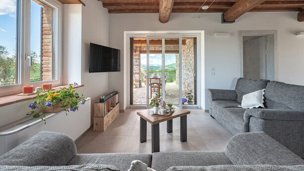 Una sala moderna con un sofá gris, una mesa de centro de madera y un televisor montado en la pared. Hay un balcón con un muro de piedra y vistas al campo.