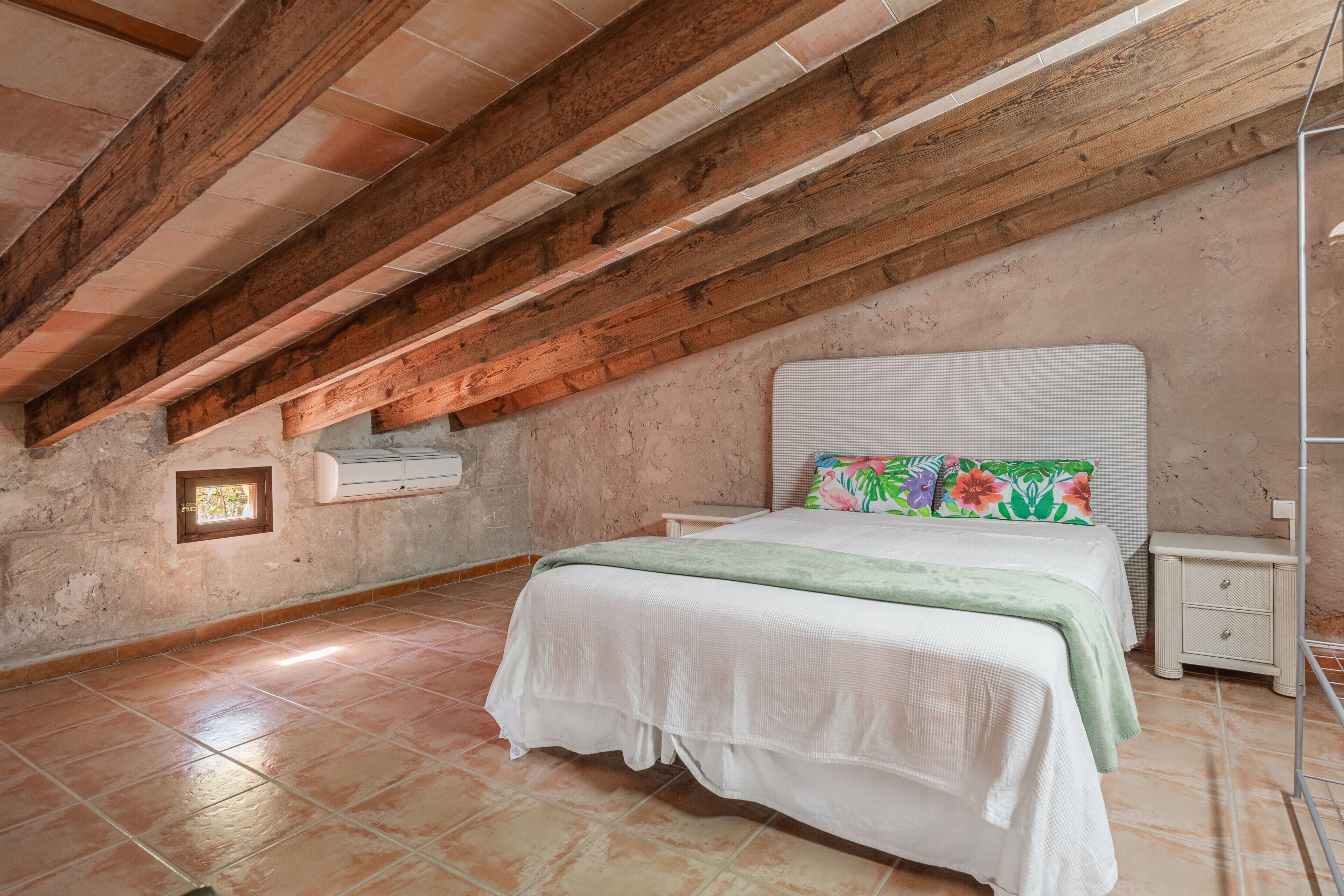 A bedroom with a sloped ceiling, a bed with a floral pillow, a bedside table, and a wall-mounted air conditioner.