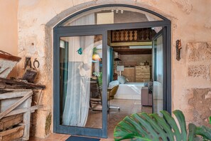 Uma porta em arco que dá entrada a um quarto com cama, cadeira e mesa.