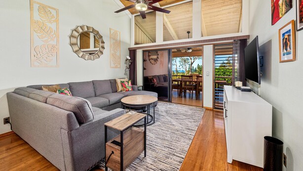 A modern living room with a gray sectional sofa, a round coffee table, and a ceiling fan. There is a TV mounted on the wall and a staircase leading to an upper level. - Summer Deal Waikoloa 3BR Villa with Pool Hot Tub and Ocean Views (Waikoloa)