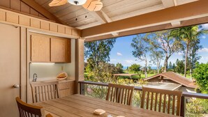 A wooden dining table with chairs, a kitchen area with wooden cabinets, and a view of a lush garden and palm trees. - Summer Deal Waikoloa 3BR Villa with Pool Hot Tub and Ocean Views (Waikoloa)