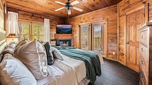 Ein Schlafzimmer einer Holzhütte mit einem Bett, einem Deckenventilator, einem Kamin und einem großen Fenster mit Rollläden.