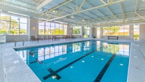 Une piscine intérieure aux parois en verre transparent, un plafond haut avec des poutres métalliques et un espace détente avec des chaises et un banc.