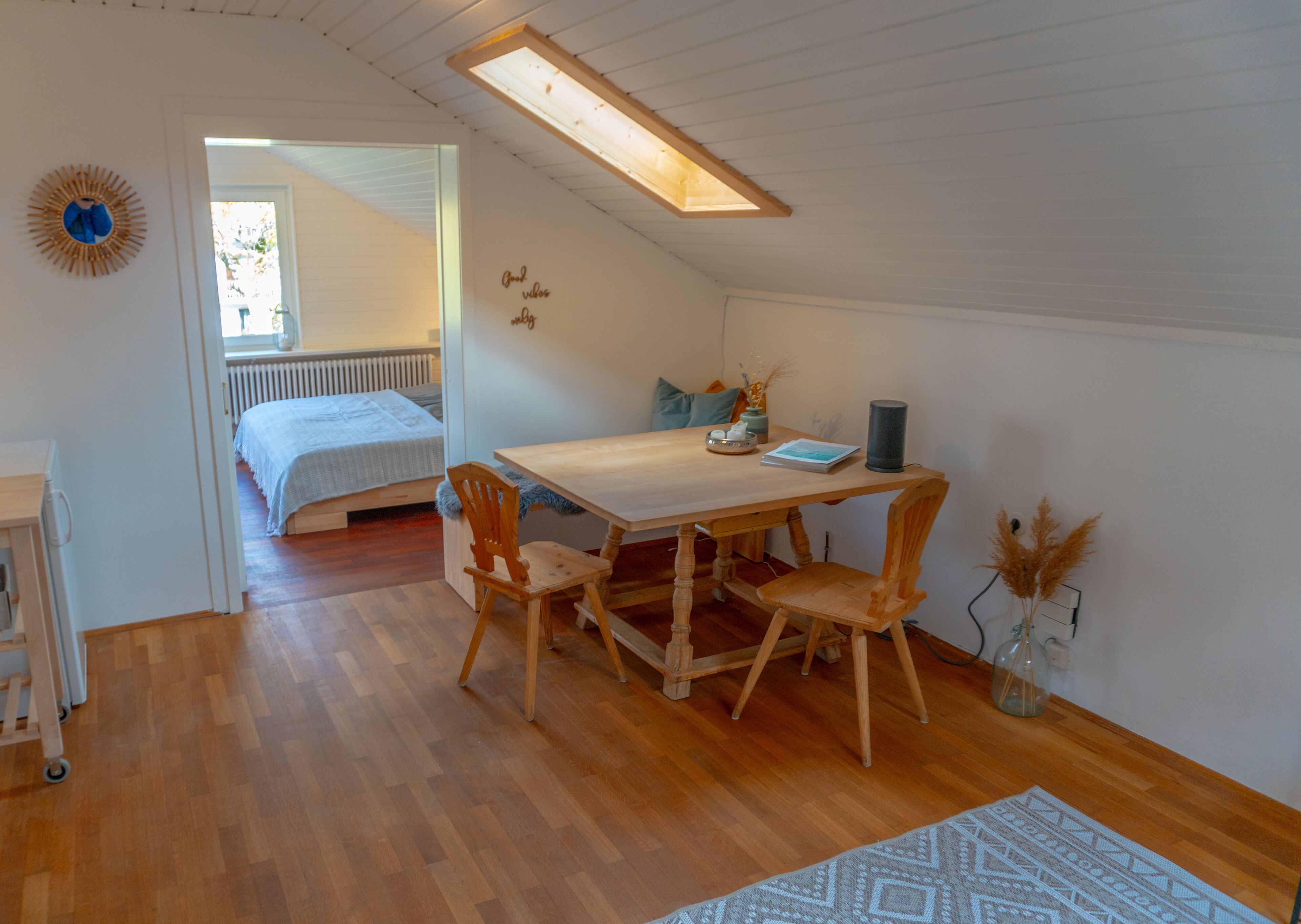 Une pièce chaleureuse et bien éclairée, avec une table à manger en bois, deux chaises, une petite table d’appoint et un lit doté d’une tête de lit blanche.