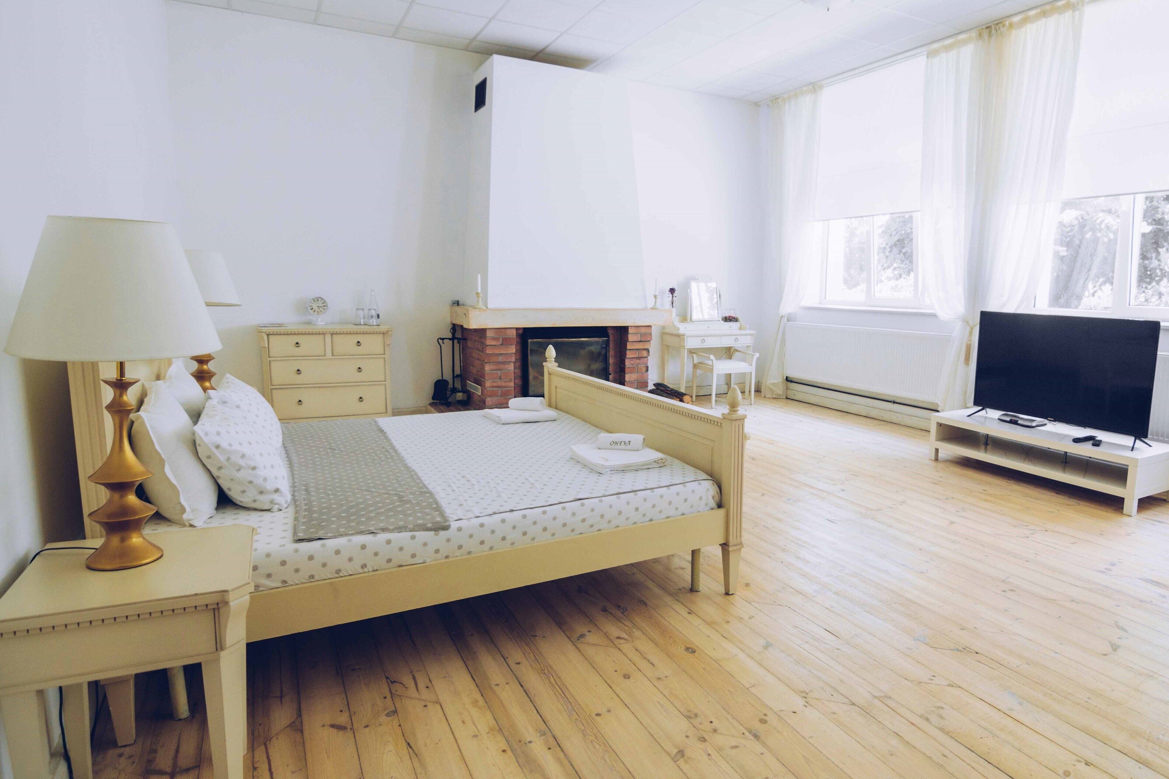 Un dormitorio con cama, mesita de noche, lámpara, cómoda, chimenea, mueble para televisión y ventanas amplias.