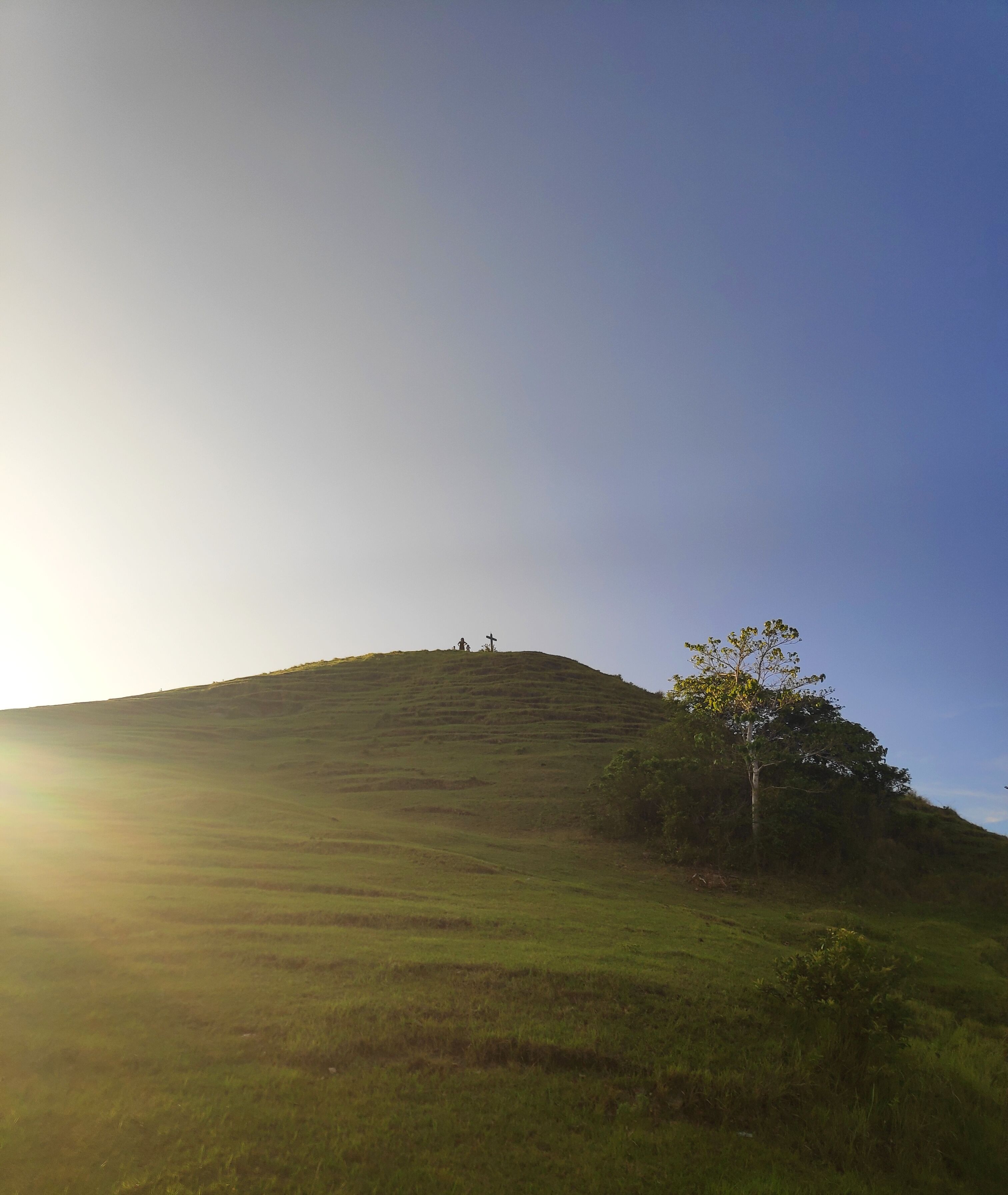 A grassy hill with a cross and two figures at the top, and a single tree on the right.