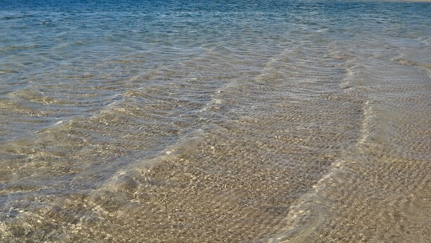 Una spiaggia con acque cristalline e poco profonde, dune sabbiose in lontananza e un cielo azzurro intenso.