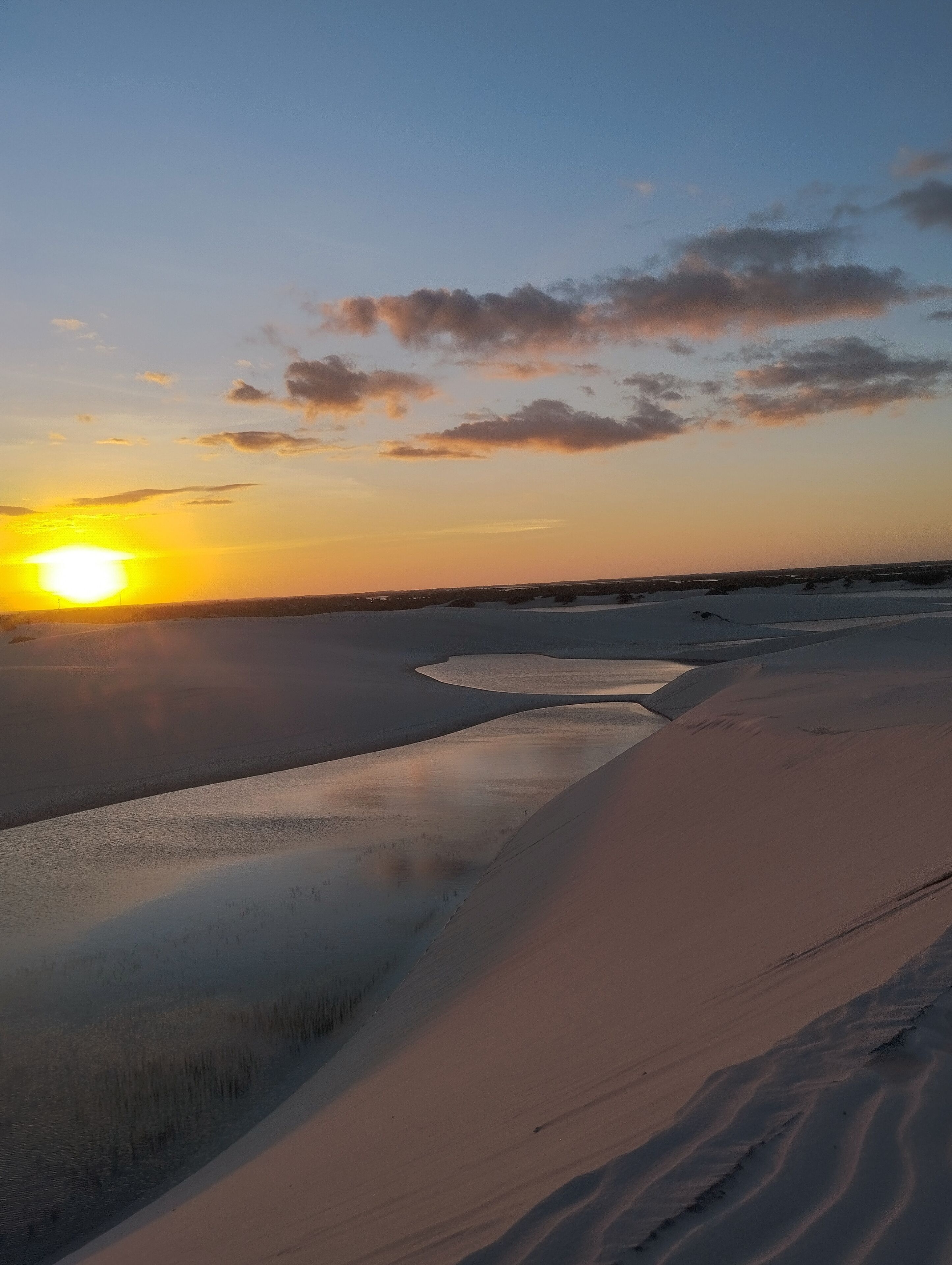 A desert landscape with sand dunes and a body of water reflecting the sunset.