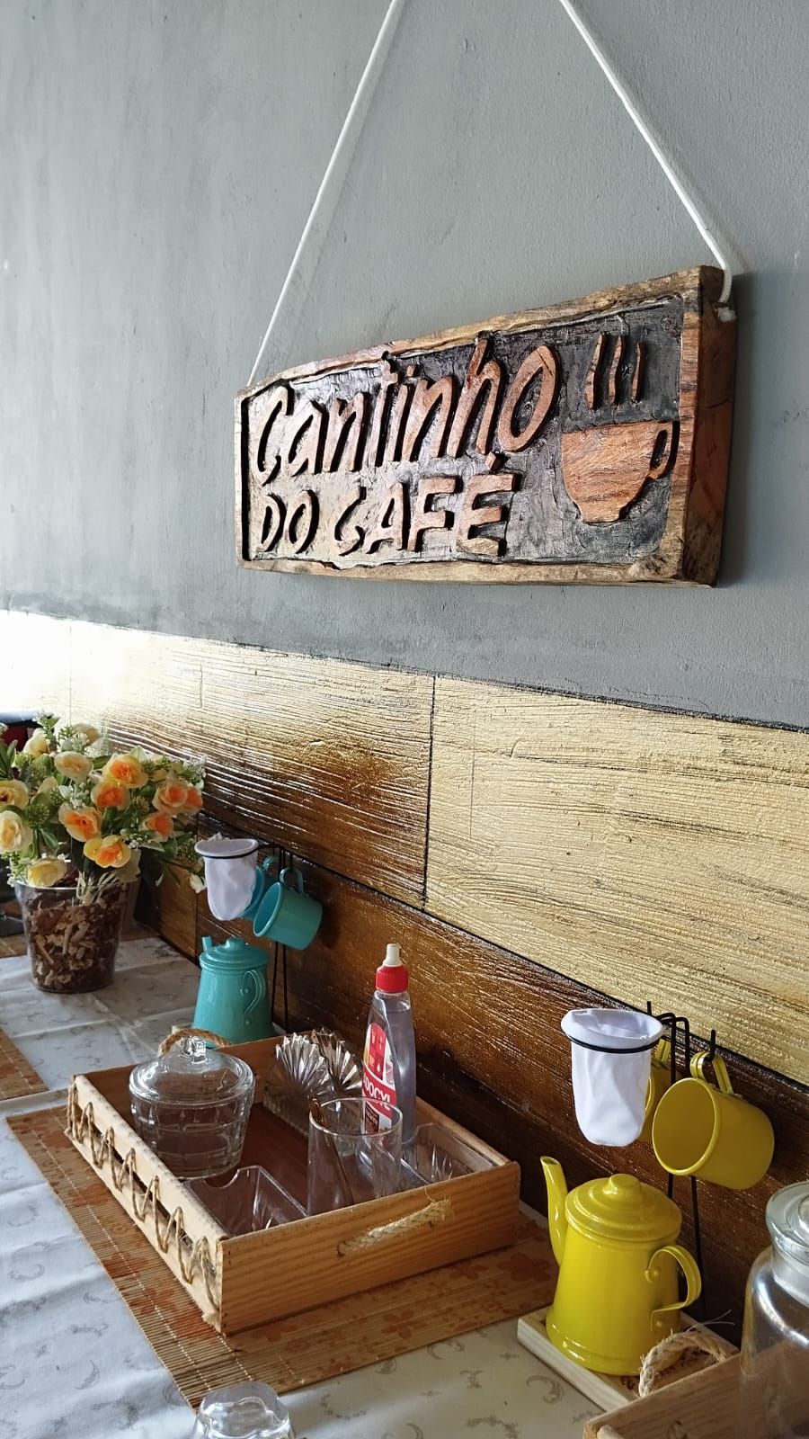Een aanrecht met een houten bordje waarop 'Cantinho do GAFE' staat, een kleine keukenopstelling met gele en witte keukengerei, een glazen pot en een fles saus.