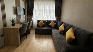 A modern living room with a dark grey sofa, wooden flooring, a desk with a laptop, and a TV mounted on the wall. - H&L Suites (Istanbul)