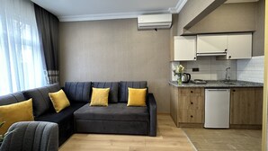 A modern living room with a dark grey sectional sofa, wooden flooring, and a kitchen area with white cabinets and a dishwasher. - H&L Suites (Istanbul)