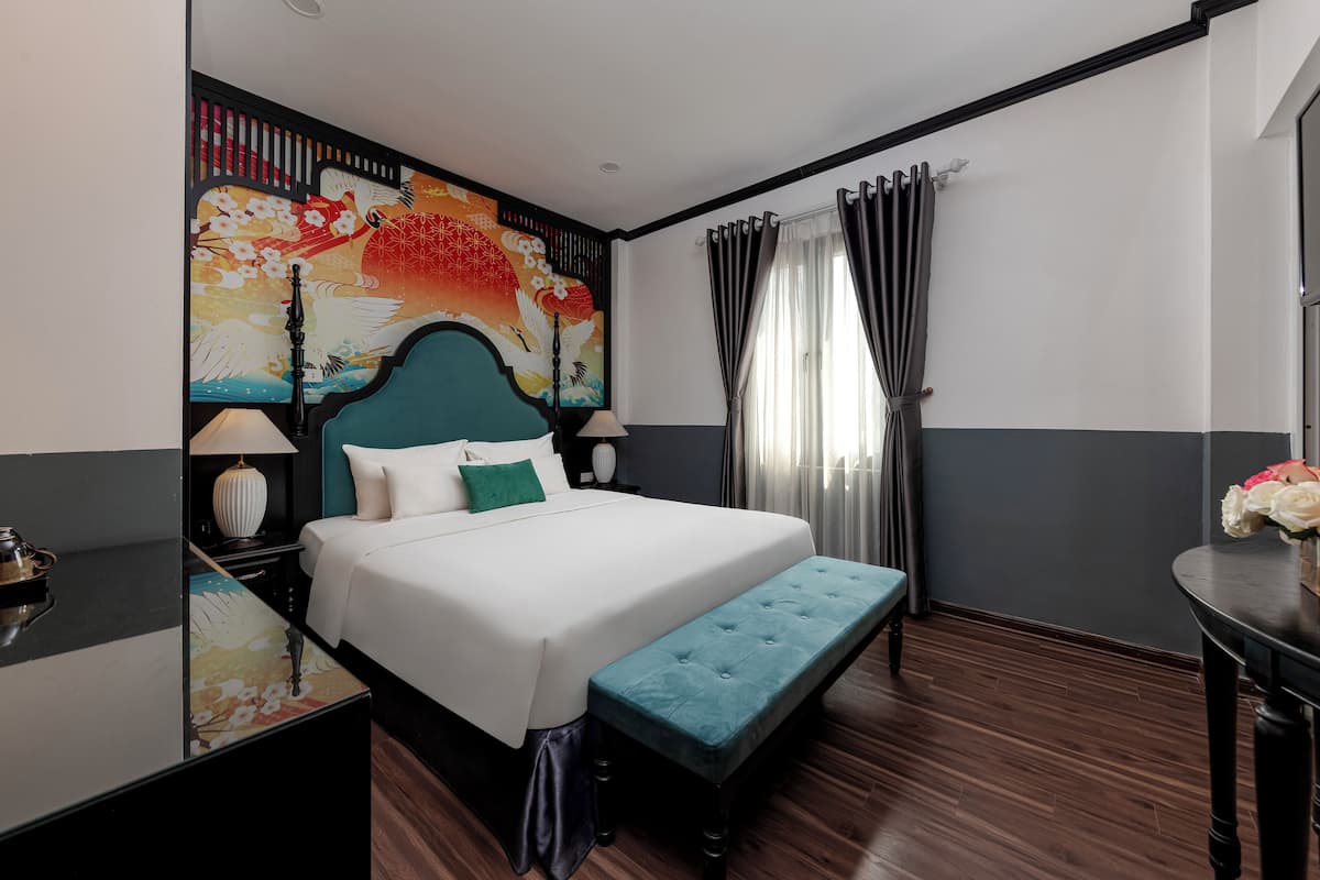 Quarto de hotel com cama grande, um banquinho, mesa de cabeceira, painel na parede e janela com cortinas.