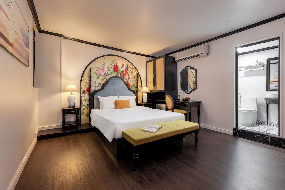 Quarto de hotel com uma cama grande, abajures, uma escrivaninha, uma cadeira e um banheiro visível por uma porta aberta.