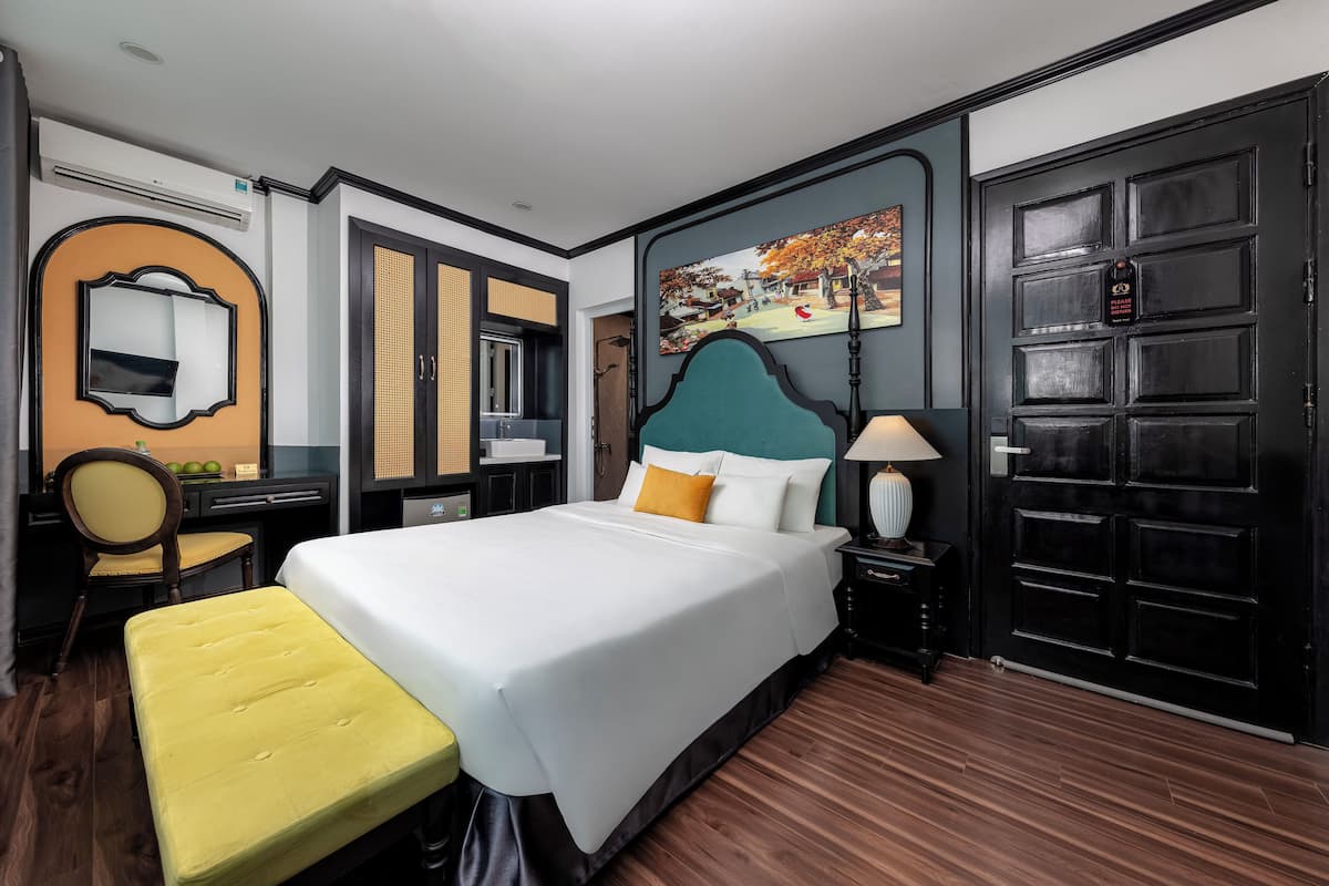 Quarto de hotel com uma cama grande, um banco, uma escrivaninha, uma cadeira, um espelho e uma porta com uma imagem.