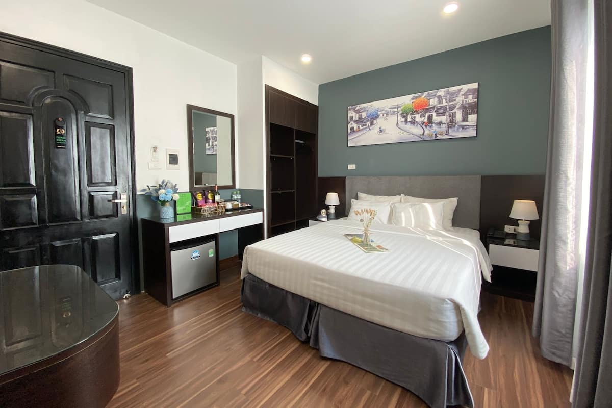 Quarto de hotel com cama, mesas de cabeceira, escrivaninha com espelho, frigorífico e uma grande janela com cortinas.