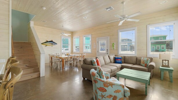 A spacious living area with a wooden staircase, a dining table with chairs, a sofa, a coffee table, and a ceiling fan. - Just Wanna Have Sun, Game Room, Pool & Beach (Port Aransas)