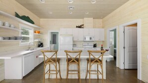 A modern kitchen with white cabinetry, a central island, and three wooden bar stools. - Just Wanna Have Sun, Game Room, Pool & Beach (Port Aransas)