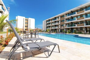A swimming pool area with lounge chairs and a dining table, surrounded by modern buildings.