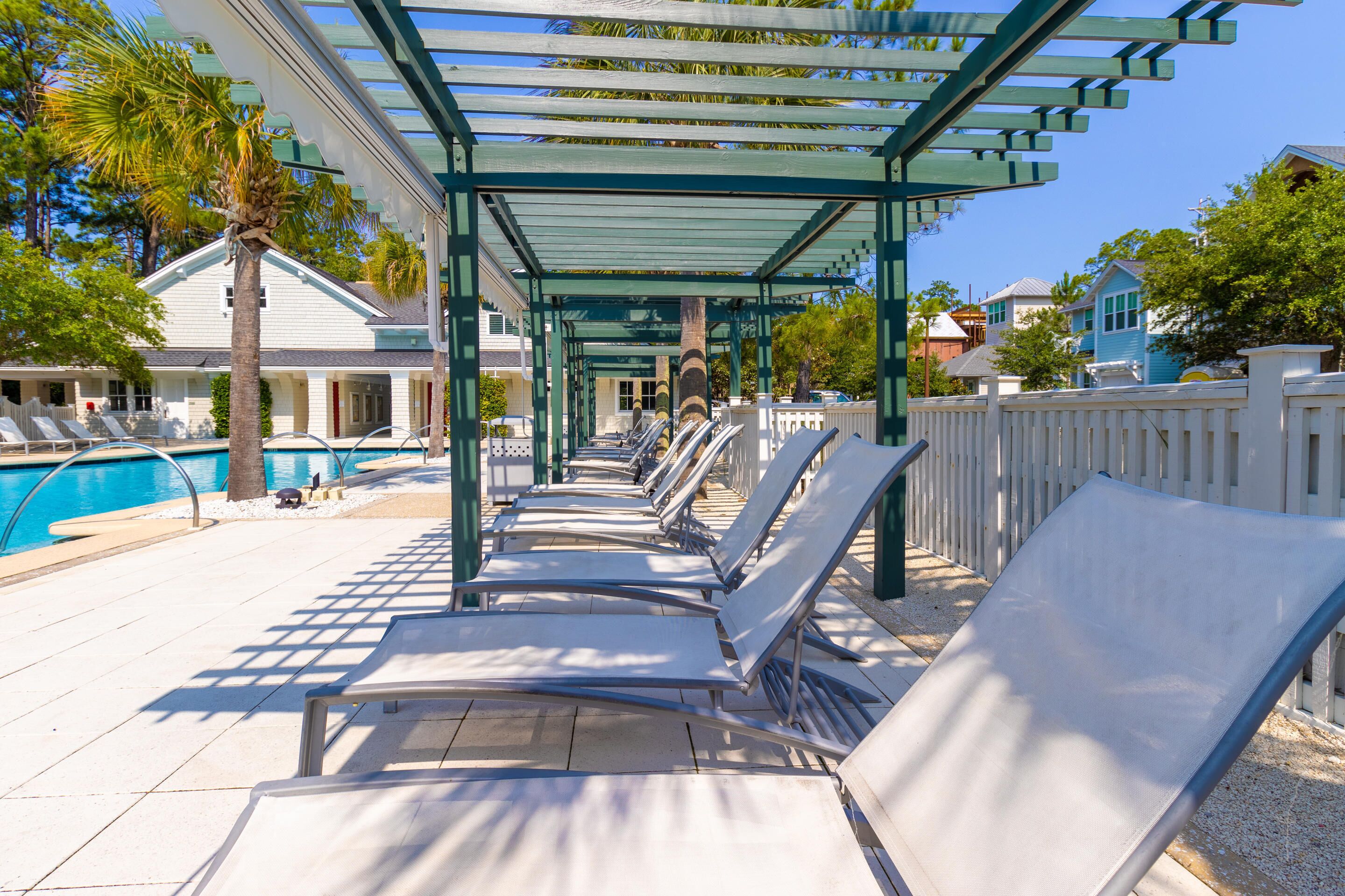 A poolside area with sun loungers under a pergola, a pool, and a white fence.