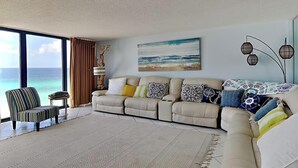 Una sala de estar con un ventanal grande con vista al océano, un sofá color beige, un sillón a rayas y una alfombra estampada.