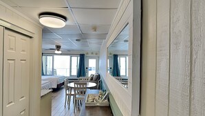 A narrow corridor with a bed, a table, and chairs, leading to a room with a large window and curtains. - Continental #512 | Private Balcony and Gulf Side Pool (Panama City Beach)