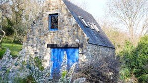 Un edificio de piedra con una puerta azul y un ventanal pequeño, rodeado de árboles y vegetación.