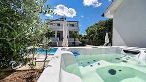 Un jacuzzi con una piscina al fondo, rodeado de follaje y un edificio.