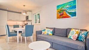 Una sala moderna con un sofá gris, almohadones coloridos, una mesa de centro blanca y un cuadro con barcos en la pared.