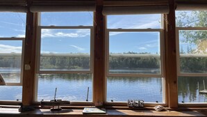 A cabin with large windows overlooking a lake, a wooden desk with a laptop, and a small decorative item on the windowsill.