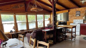 A cabin with a wooden interior, large windows overlooking a lake, a dining area with a table and chairs, a kitchen area with a stove and refrigerator, and a cozy seating area with a couch and pillows.