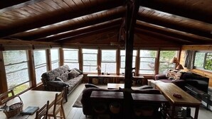 A cozy cabin interior with a wooden ceiling, large windows, a sofa, a dining table, and a fireplace.