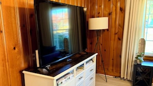 A room with wooden paneling, a flat-screen TV on a white dresser, a floor lamp, and a window with curtains. - 3 bedroom cozy yet spacious home w/ in-ground pool (Richmond)