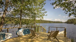 Une terrasse en bois avec deux chaises longues et un kayak, donnant sur un lac paisible bordé d’arbres.