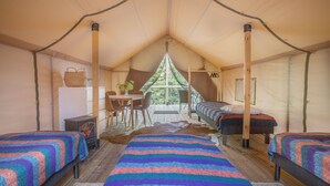 Un hébergement sous tente comprenant deux lits, une table, des chaises, un petit chauffage et une fenêtre donnant sur un paysage verdoyant.