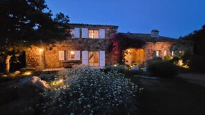 Edificio de piedra con múltiples ventanas y puertas, rodeado de un jardín con flores blancas y rocas.