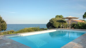 Una piscina con vistas al mar, rodeada de vegetación y un patio con sillas y una sombrilla.