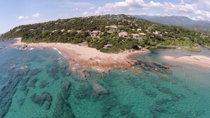 Vista aérea de una zona costera con aguas turquesas cristalinas, una playa de arena y una colina con edificios dispersos.