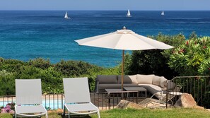 A poolside area with lounge chairs and a table, overlooking a clear blue sea with sailboats. -  Exclusive granite villa DIRECTLY on the beach, sea and mountain panorama, pool (Zonza)