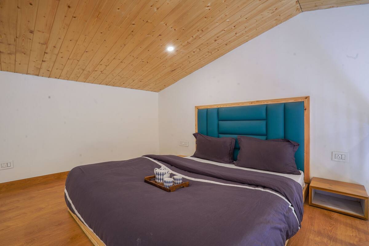 A bedroom with a wooden ceiling, a bed with a teal headboard, and a tray with cups on the bed.