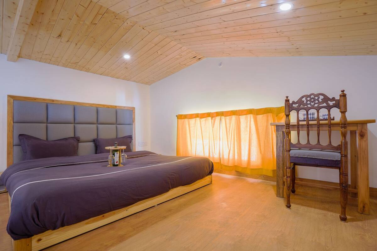 A bedroom with a wooden ceiling, a bed with a headboard, a wooden chair, and a window with curtains.