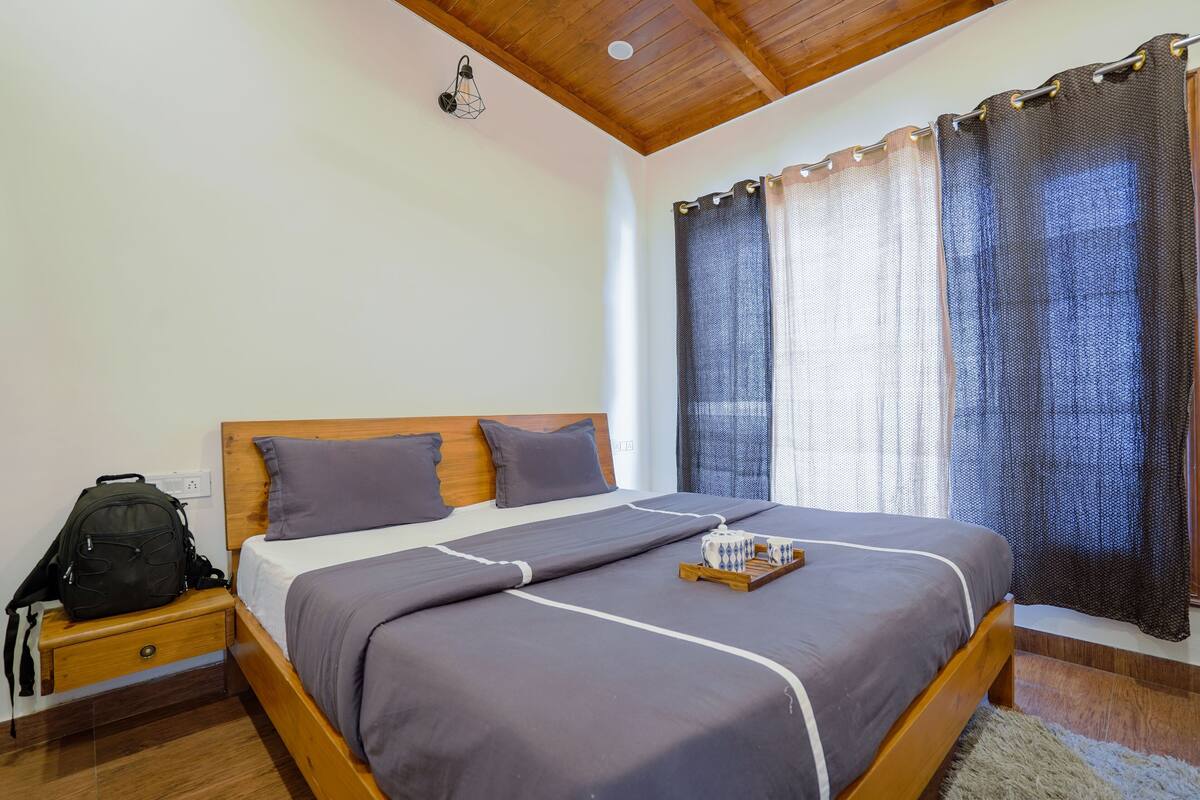 A neatly made bed with a wooden headboard, a bedside table with a backpack, and a window with curtains.