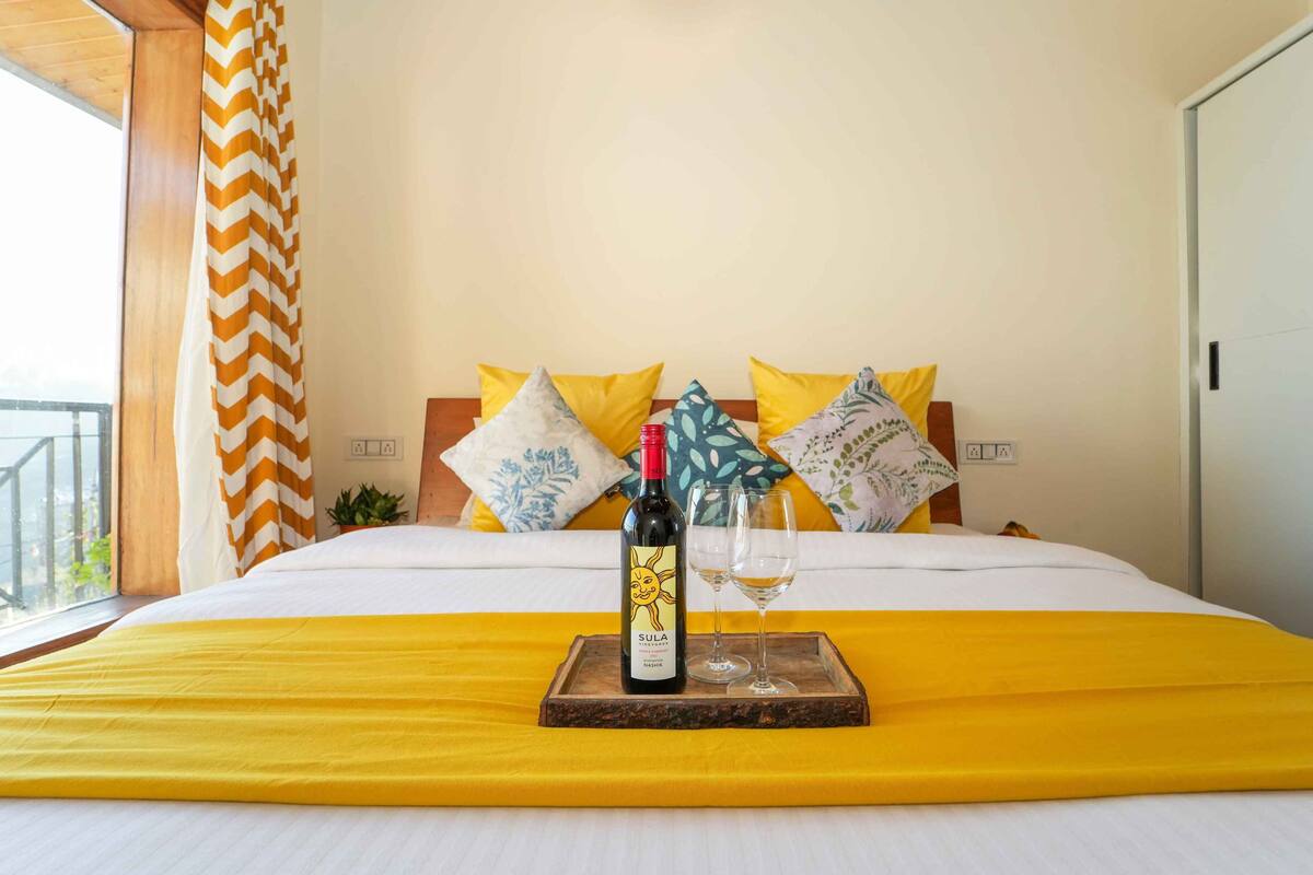 A bed with a yellow bedspread, a bottle of Sula wine, and two glasses on a wooden tray.